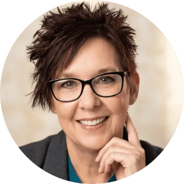 Smiling woman with short auburn hair, glasses, and gray blazer. Confident, approachable headshot with hand to face.