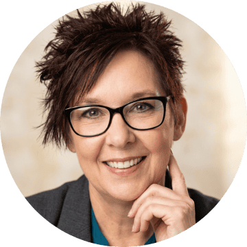 Smiling woman with short auburn hair, glasses, and gray blazer. Confident, approachable headshot with hand to face.